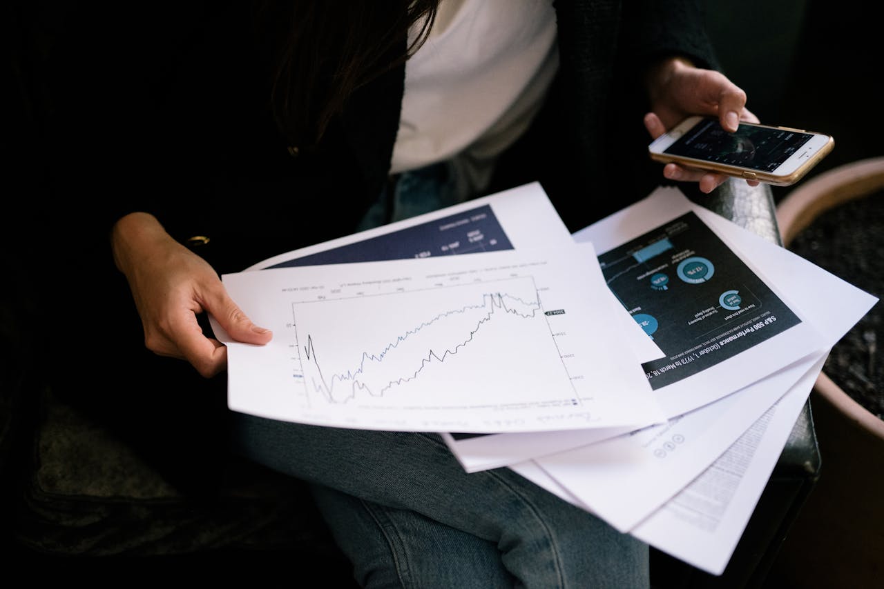 creative-03 A person reviews stock market graphs on paper and smartphone for business insights.